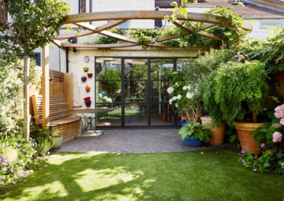 Back garden design in Frankfurt Road, 1 View from back of garden towards house. Lawn, side beds and pots with flowers, and patio with storage bench and table beneath pergola.