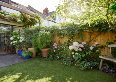 Back garden design in Frankfurt Road, 3 Lawn, border with flowering plants, and patio pot plants.